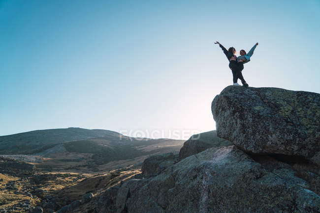 Paesaggio paesaggistico della valle delle montagne con due ragazze abbracciate con le braccia sollevate che posano sul masso — Foto stock