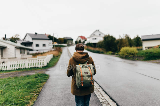 Rear view of woman with backpack walking down village street on dull day. — Stock Photo
