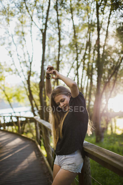 Jovem loira apoiada em trilhos de ponte de madeira e posando para câmera no dia ensolarado . — Fotografia de Stock