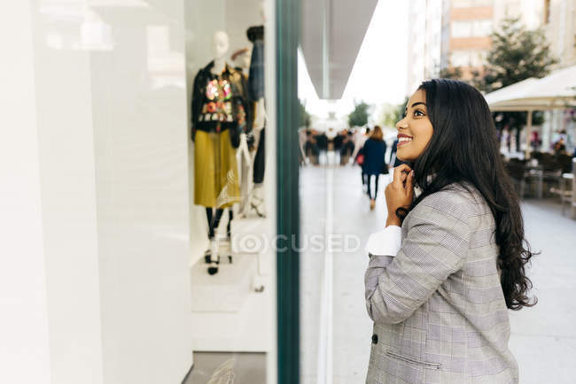 Vista lateral de la mujer sonriente con chaqueta mirando al escaparate - foto de stock