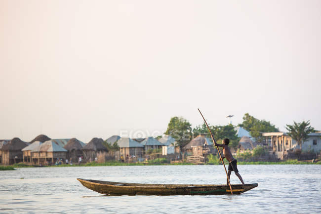 BENIN, AFRIQUE - 30 AOÛT 2017 : Vue latérale d'un garçon conduisant un bateau avec un bâton sur un lac sur fond de village africain . — Photo de stock