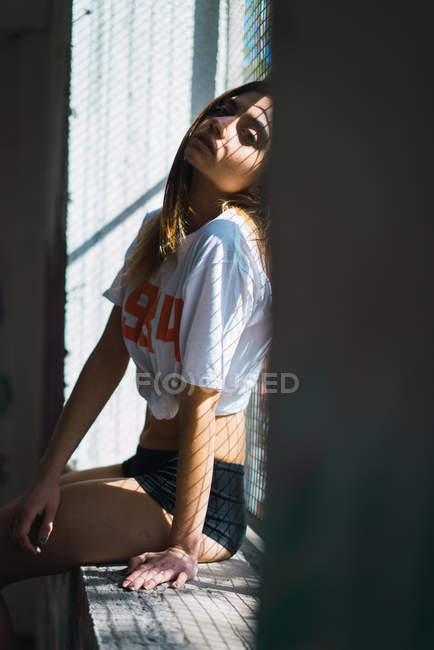 Портрет чуттєвої дівчини в футболці позує біля підвіконня з сіткою — стокове фото