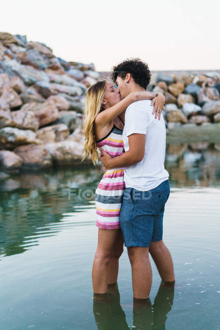 Seitenansicht eines sich umarmenden Pärchens, das im Wasser steht und sich vor dem Hintergrund von Küstensteinen küsst — Stockfoto