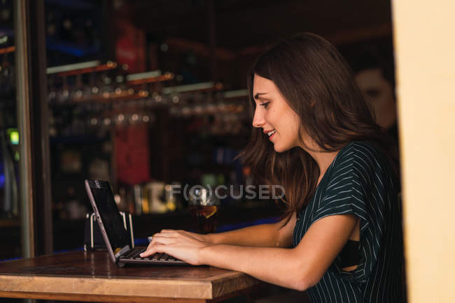 Vista lateral de la mujer sonriente escribiendo en el ordenador portátil en la cafetería - foto de stock