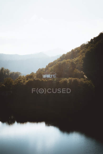 Vue lointaine sur petite maison au bord du lac avec des montagnes verdoyantes dans le brouillard en arrière-plan  . — Photo de stock