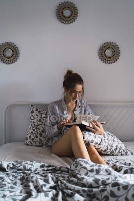 Portrait of brunette girl sitting on bed in shirt and reading book — Stock Photo