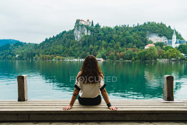 Rückansicht einer brünetten Frau, die auf einem Steg sitzt und auf Hügel über dem See blickt — Stockfoto