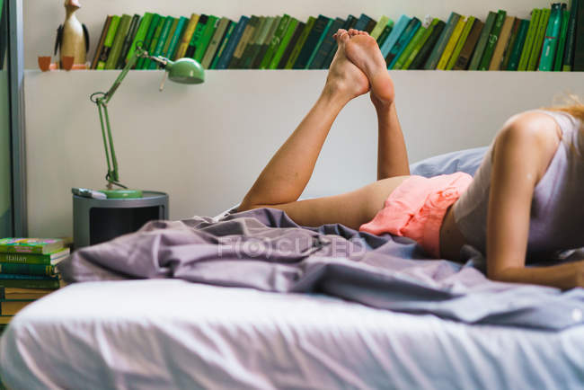 Crop donna sdraiata sul letto sopra scaffale libro — Foto stock