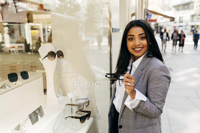 Bella donna elegante con occhiali da sole guardando la fotocamera vicino alla finestra del negozio . — Foto stock