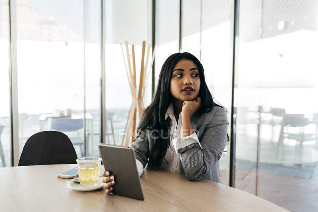 Elegante mujer de negocios con tableta sentada en la mesa y mirando a un lado - foto de stock