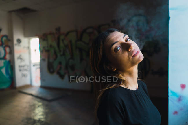 Porträt eines brünetten Mädchens, das am Fenster raucht, verlassener Raum mit Graffiti an Wänden. — Stockfoto