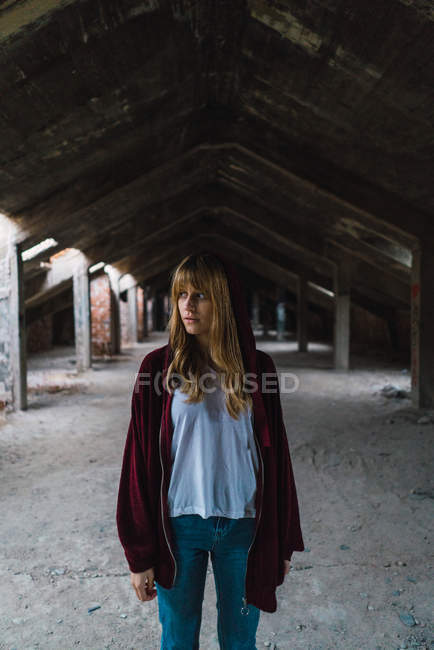 Chica en chaqueta con capucha roja posando en un edificio abandonado y mirando hacia otro lado - foto de stock