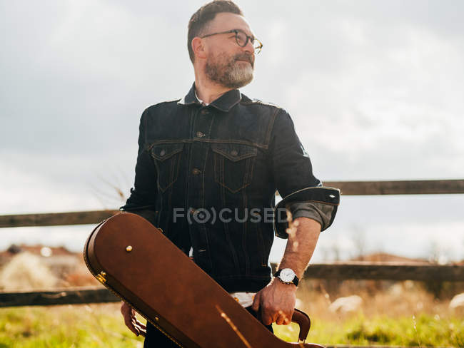 Ritratto di uomo barbuto grigio in posa con custodia per chitarra alla recinzione di campagna e distogliendo lo sguardo — Foto stock