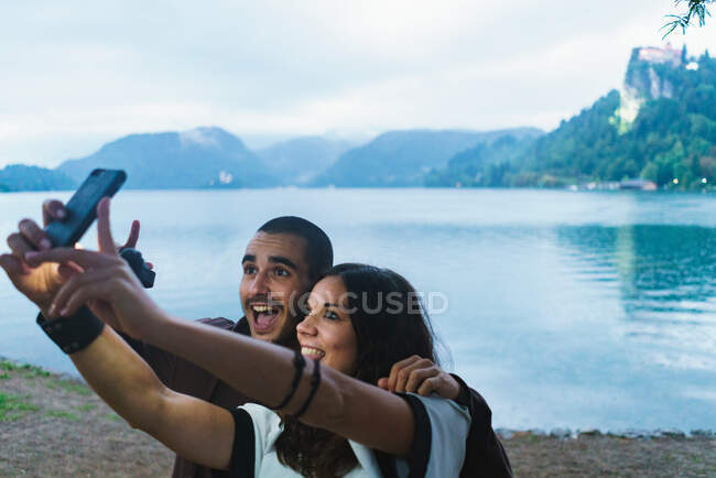 Casal alegre se divertindo e tirando selfie com smartphone na margem do lago juntos. — Fotografia de Stock