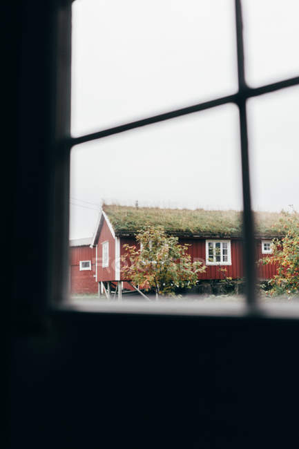 Vue de la maison rouge avec toit recouvert d'herbe depuis la fenêtre de la pièce sombre . — Photo de stock