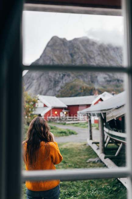 Rear view of woman in orange jumper standing near boat shed and enjoying view of mountain village — Stock Photo
