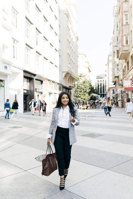 Улыбающаяся элегантная деловая женщина с сумочкой, гуляющая по улице — стоковое фото