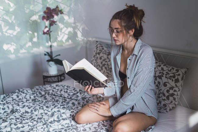 Girl in eyeglasses sitting on bed and reading book — Stock Photo