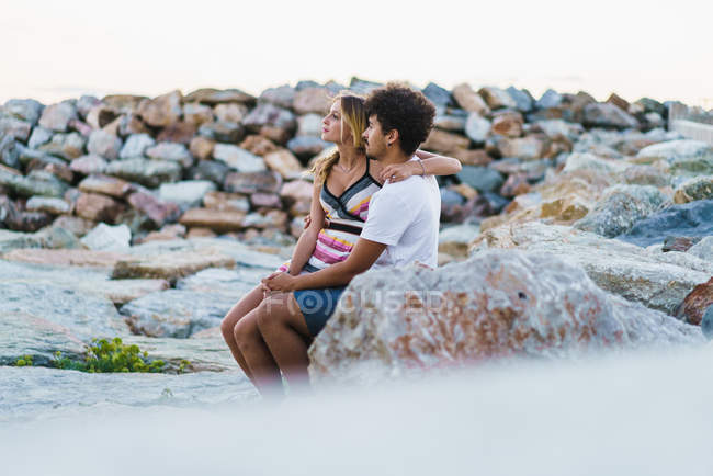 Jeune couple embrassant assis sur des rochers sur le littoral et regardant loin . — Photo de stock