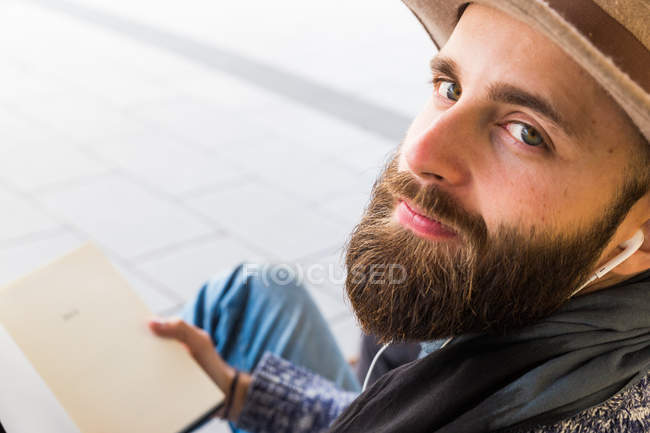 Бородатый мужчина слушает музыку с книгой в руках и смотрит в камеру — стоковое фото