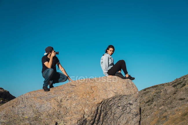 Vista lateral del fotógrafo tomando la foto de la niña sentada en el borde de la roca - foto de stock