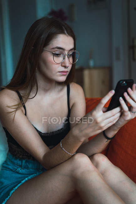 Retrato de menina morena em óculos vestindo roupa casual relaxante em casa e usando smartphone . — Fotografia de Stock