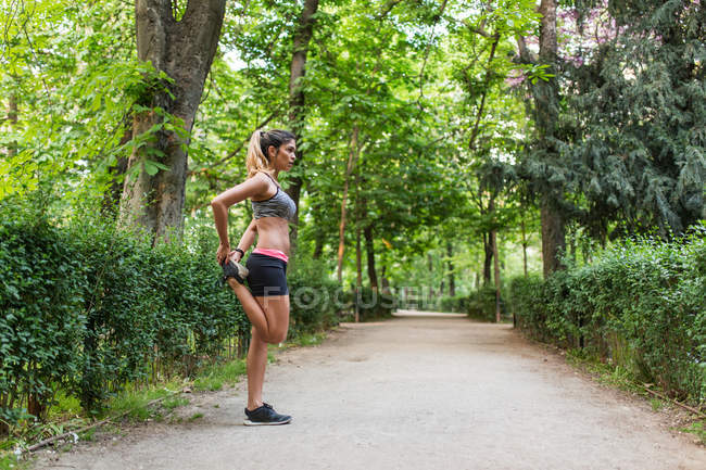 Side view of fit woman in sportswear stretching leg on pathway in green park — Stock Photo