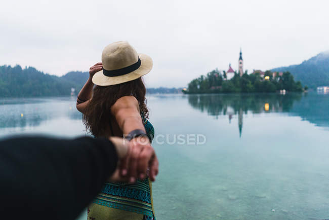 Rückansicht eines brünetten Mädchens mit Strohhut folgt mir gestikulierend am Bergsee — Stockfoto