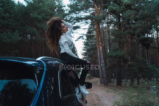 Morena mulher sentada em cima do carro estacionado no lado da estrada na floresta — Fotografia de Stock