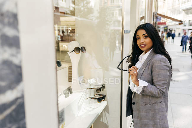 Портрет елегантної жінки позує біля вікна магазину з окулярами в руці і дивиться на камеру — стокове фото