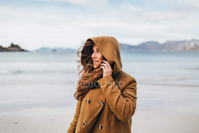 Smiling brunette woman wearing coat hood posing over mountain lake and looking away — Stock Photo