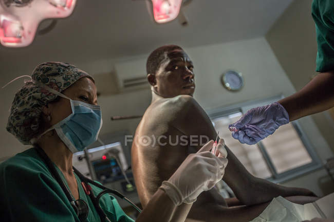 BENIN, AFRICA - AUGUST 31, 2017: Black man sitting in hospital and looking over shoulder at camera while staff making  treatment. — Stock Photo