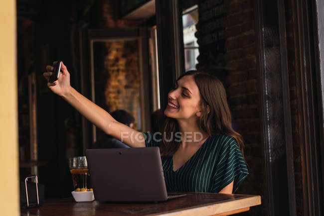 Retrato de mulher sentada à mesa do café com laptop e tomando selfie — Fotografia de Stock