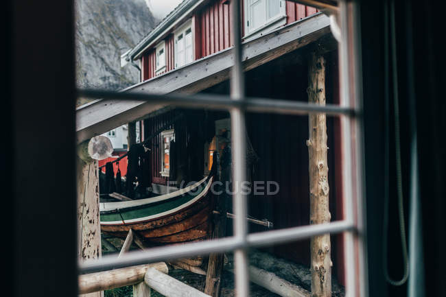 View through window of old fishing boat  standing under roof beside red house — Stock Photo