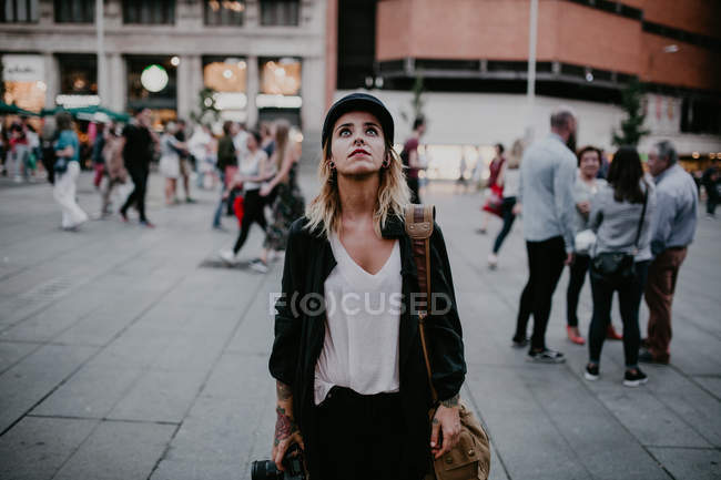 Mulher com câmera nas mãos em pé na rua da cidade e olhando para cima — Fotografia de Stock
