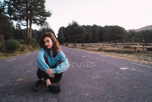 Attractive brunette woman sitting on rural road and looking at camera. — Stock Photo