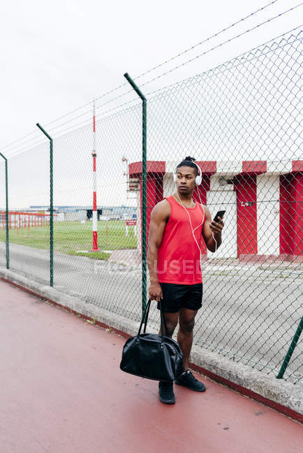 Sportler mit Kopfhörer hält Handtasche und Smartphone in den Händen und schaut weg — Stockfoto