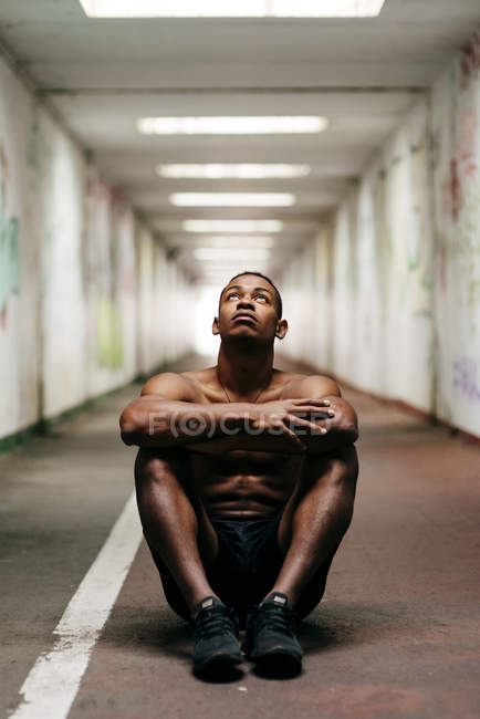 Спортсмен без рубашки сидит на полу в подземном переходе и смотрит вверх — стоковое фото