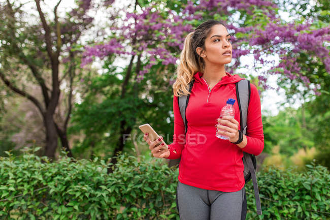 Chica atlética posando con teléfono inteligente y botella de agua en las manos en el parque - foto de stock