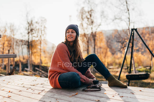 Brunette woman sitting with camera on wooden terrace in forest and looking away — Stock Photo