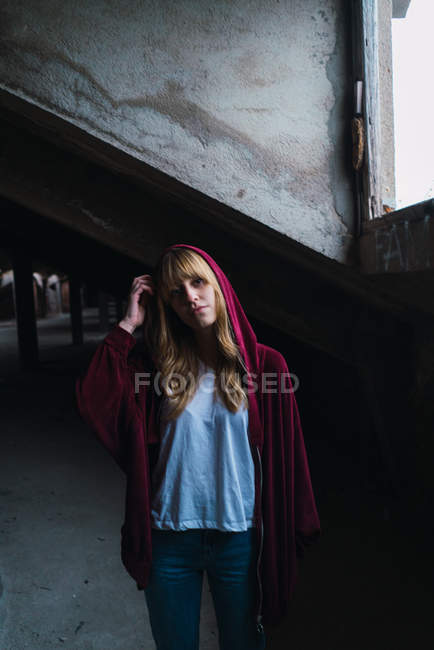 Pretty young woman in red jacket with hood standing in derelict building near window. — Stock Photo