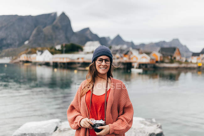 Mulher morena sorridente em óculos segurando câmera de filme nas mãos e olhando para a câmera no cais do lago — Fotografia de Stock