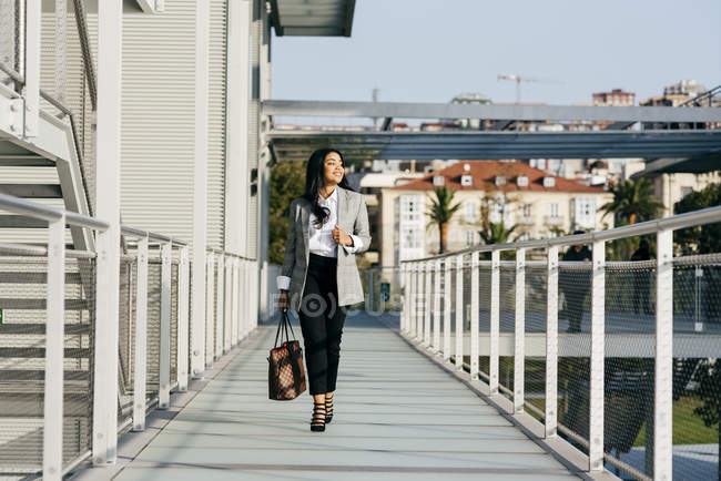 Vista frontal de la elegante mujer de negocios llevando bolso y caminando en el pasillo del balcón - foto de stock