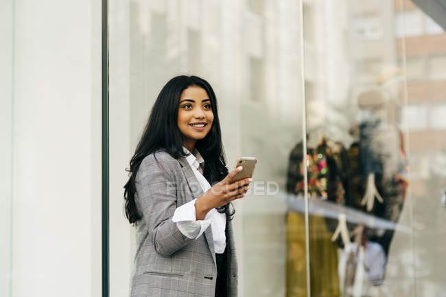 Retrato de una alegre empresaria sosteniendo un smartphone y mirando hacia otro lado cerca del escaparate - foto de stock