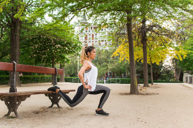 Vista lateral da menina atlética esticando as pernas no banco no parque — Fotografia de Stock