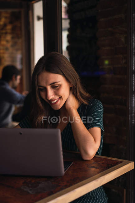 Portrait of smiling woman touching neck while browsing laptop computer in cafe. — Stock Photo