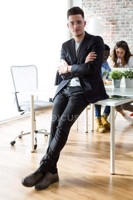 Портрет молодого бизнесмена, склонившегося за столом и смотрящего в камеру на рабочем месте офиса — стоковое фото