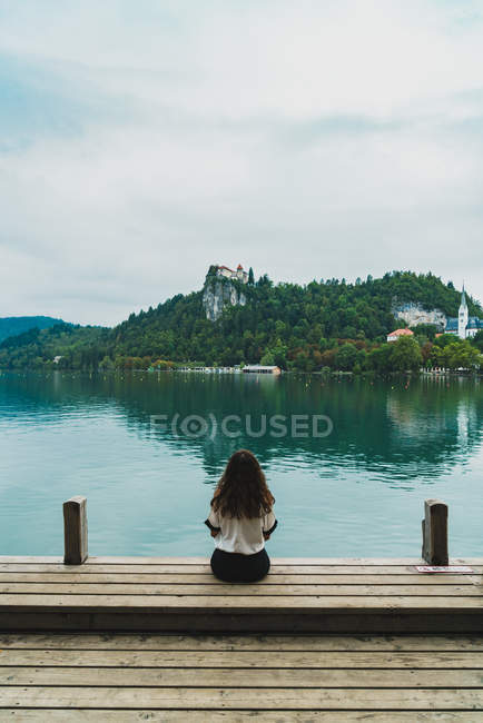 Visão traseira da mulher sentada no cais de madeira no lago e olhando para colinas na costa oposta — Fotografia de Stock