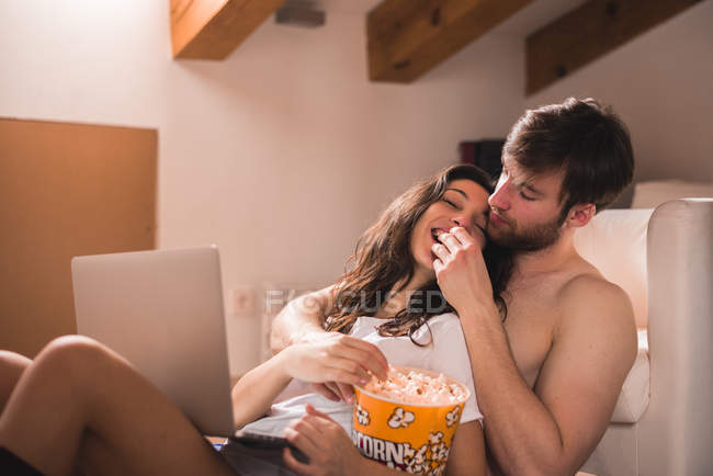 Homem alimentando namorada com pipocas enquanto navega laptop . — Fotografia de Stock