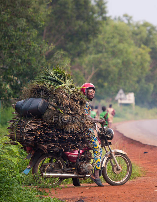BENIN, ÁFRICA - 31 de agosto de 2017: Vista lateral del hombre sentado en motocicleta con montones de heno y ramitas sobre fondo de carretera tropical . - foto de stock